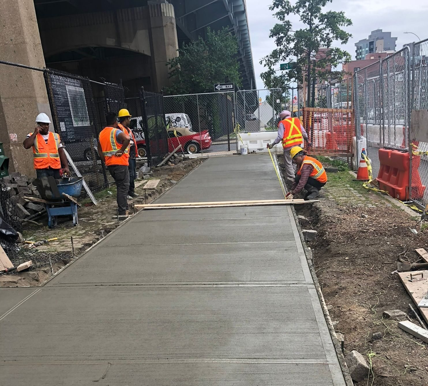Construction crew smoothing and measuring a newly poured concrete sidewalk near an underpass in NYC.