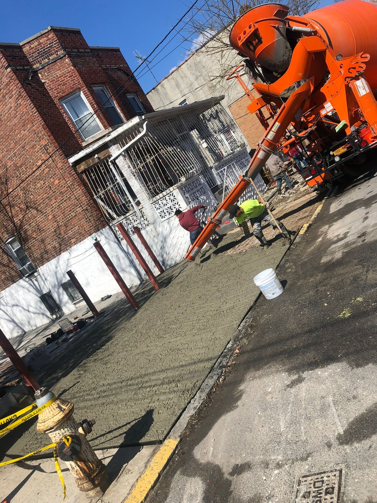 Construction crew pouring and leveling concrete beside a brick residential building using an orange cement truck in NYC.