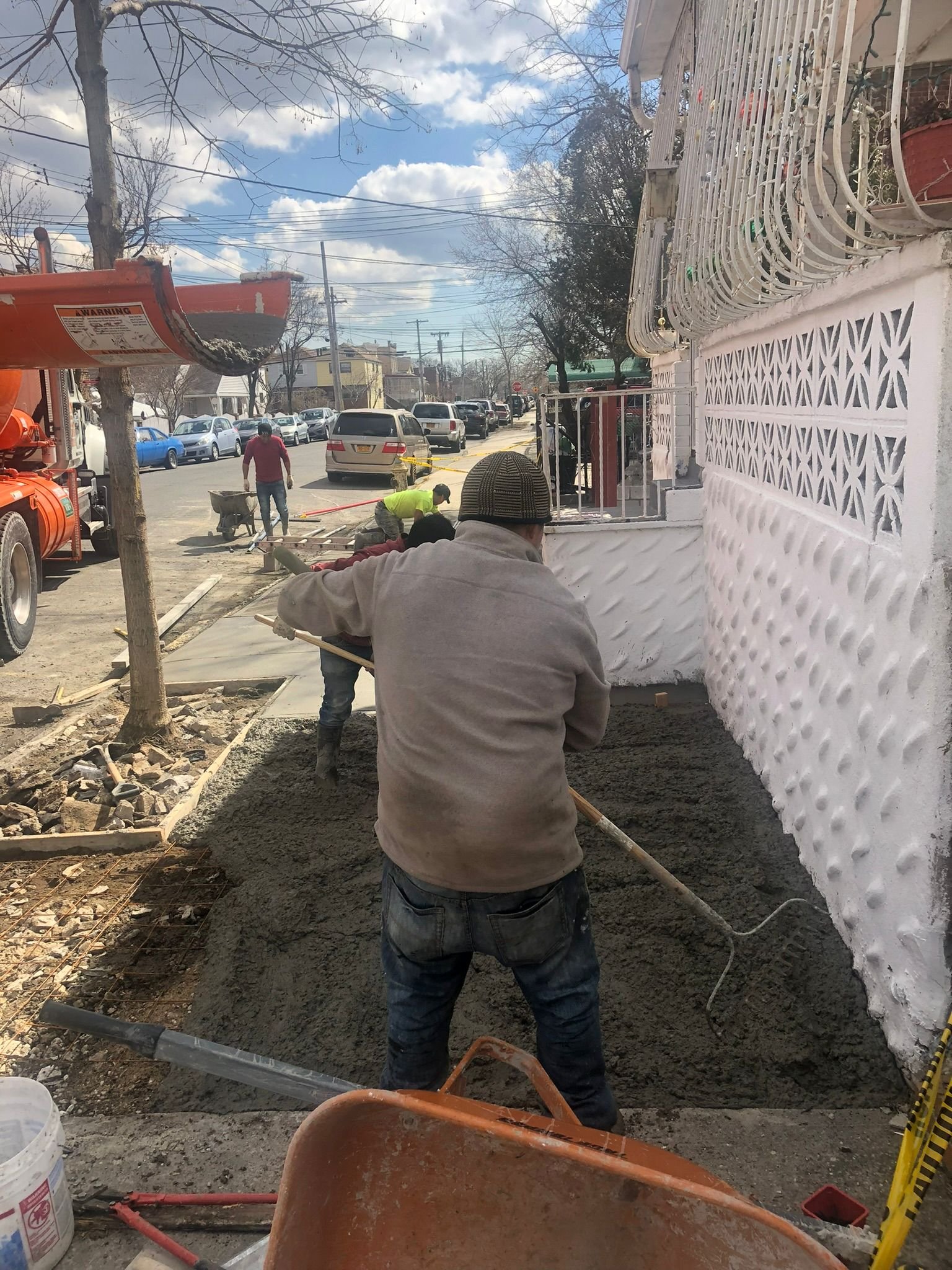 Concrete crew pouring and spreading concrete on a residential NYC street using an orange mixer truck.