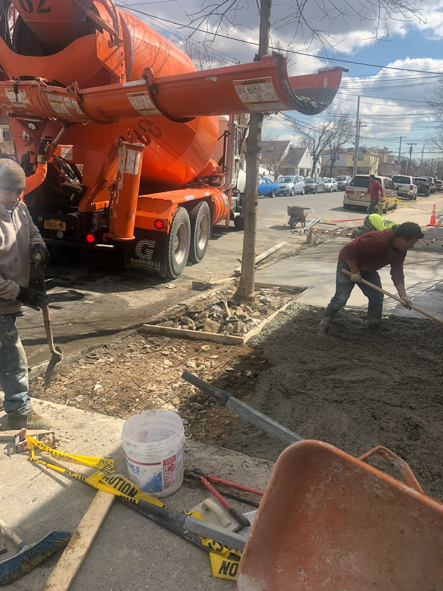 Construction workers spreading and leveling fresh concrete on a residential street beside an orange cement mixer truck in NYC.